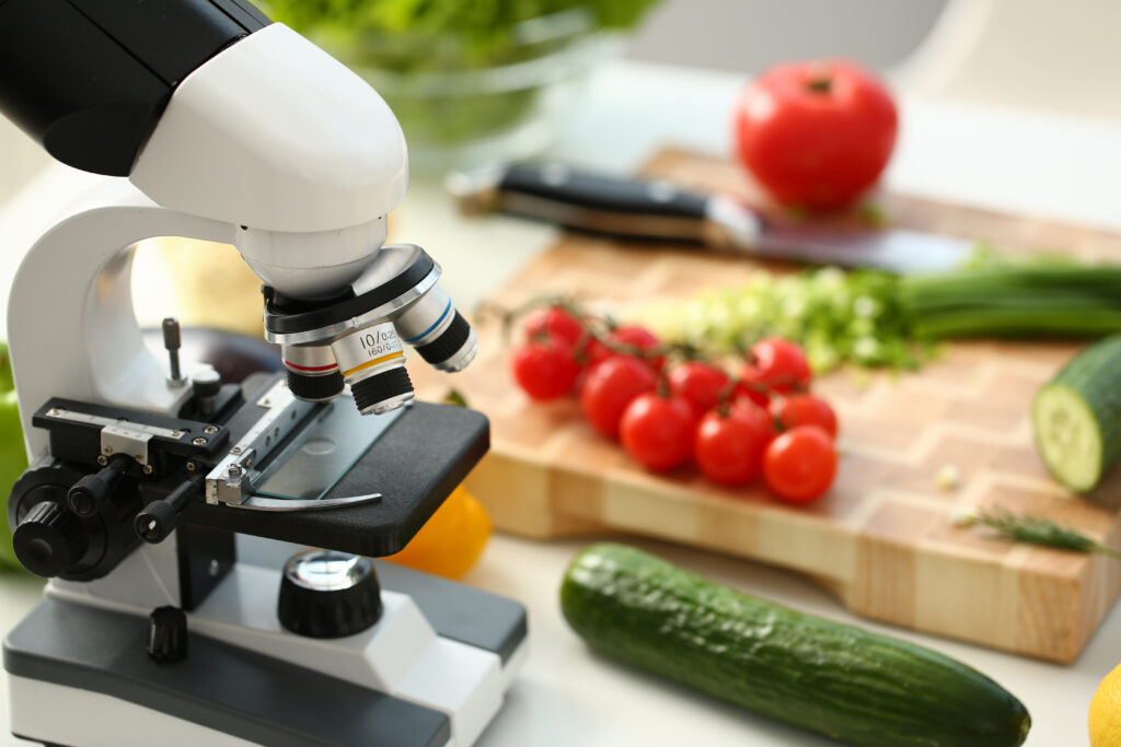 Close up of laboratory optical microscope, rope tomatoes, cucumbers, knife and wooden cutting board on desk